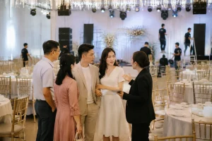 Cô dâu chú rể Việt Nam cùng người thân quan sát khu vực chuẩn bị tiệc cưới với một điều phối viên nhà hàng, thể hiện việc gom nhiều hạng mục tổ chức về một đầu mối trong quá trình