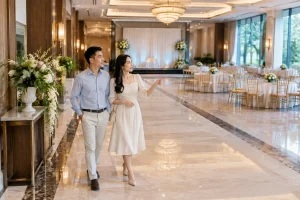 Cặp đôi Việt Nam đang tham quan sảnh tổ chức tiệc cưới trong một nhà hàng, cùng quan sát cách bố trí lối đi, khu vực đón khách và không gian tổng thể trước khi đưa ra quyết định, g
